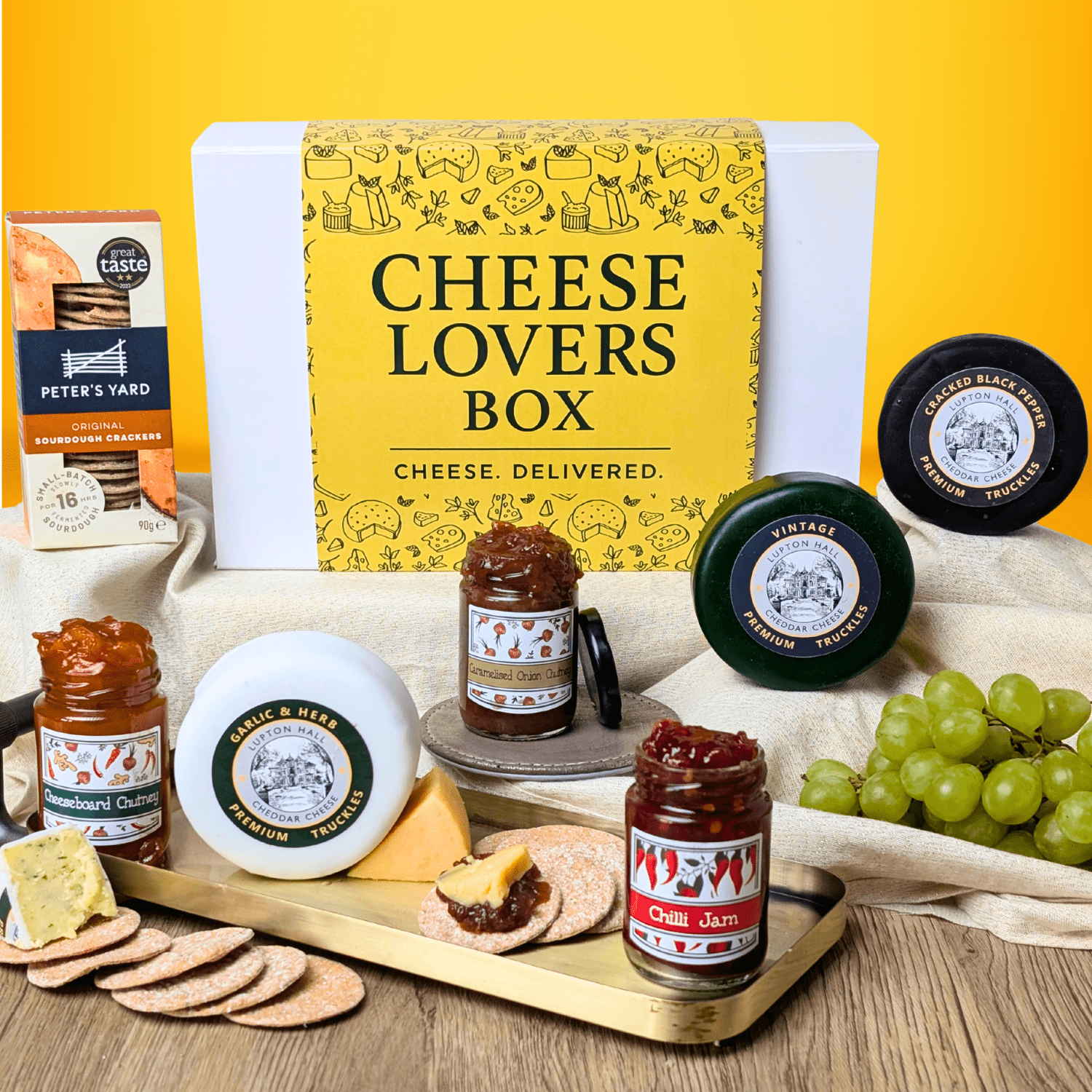 Cheese Gift Box with various cheeses, jams, and crackers on a wooden board against a yellow background.